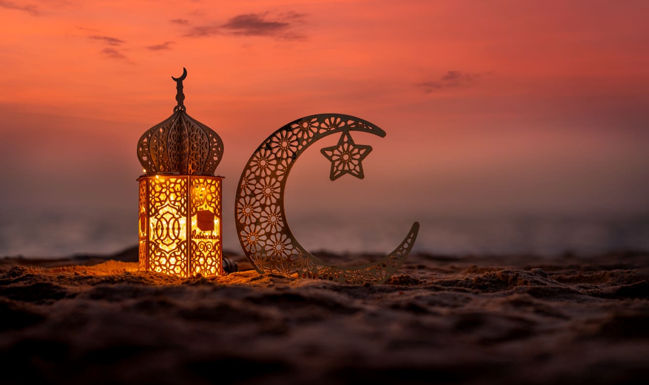 A beautifully lit Ramadan lantern and ornate crescent moon rest on the sand under a warm, glowing sunset sky.