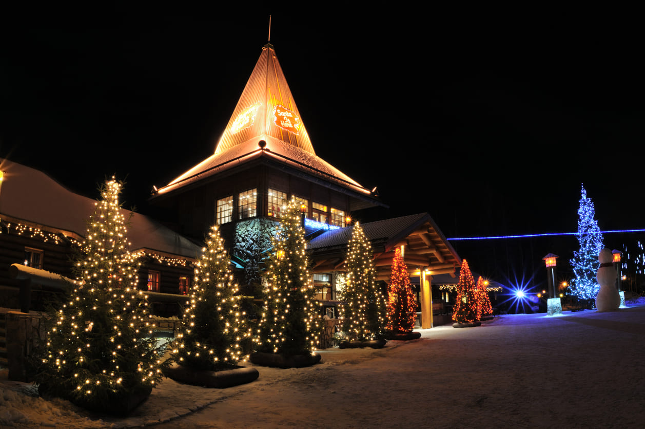 A snowy evening scene at Santa Claus Village in Rovaniemi, beautifully lit with glowing Christmas trees, festive decorations, and a towering snowman.