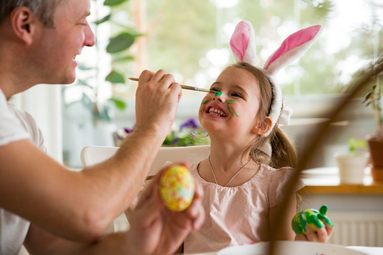 A smiling father paints his daughter's face with green whiskers as she wears bunny ears, enjoying Easter crafts in a cozy Finnish home.