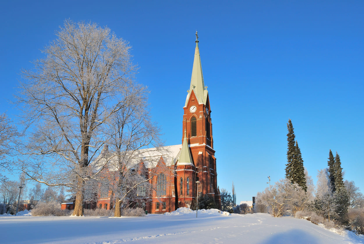 A tall red-brick church surrounded by snow-covered trees under a bright blue sky, capturing the tranquil beauty of a Finnish winter morning.