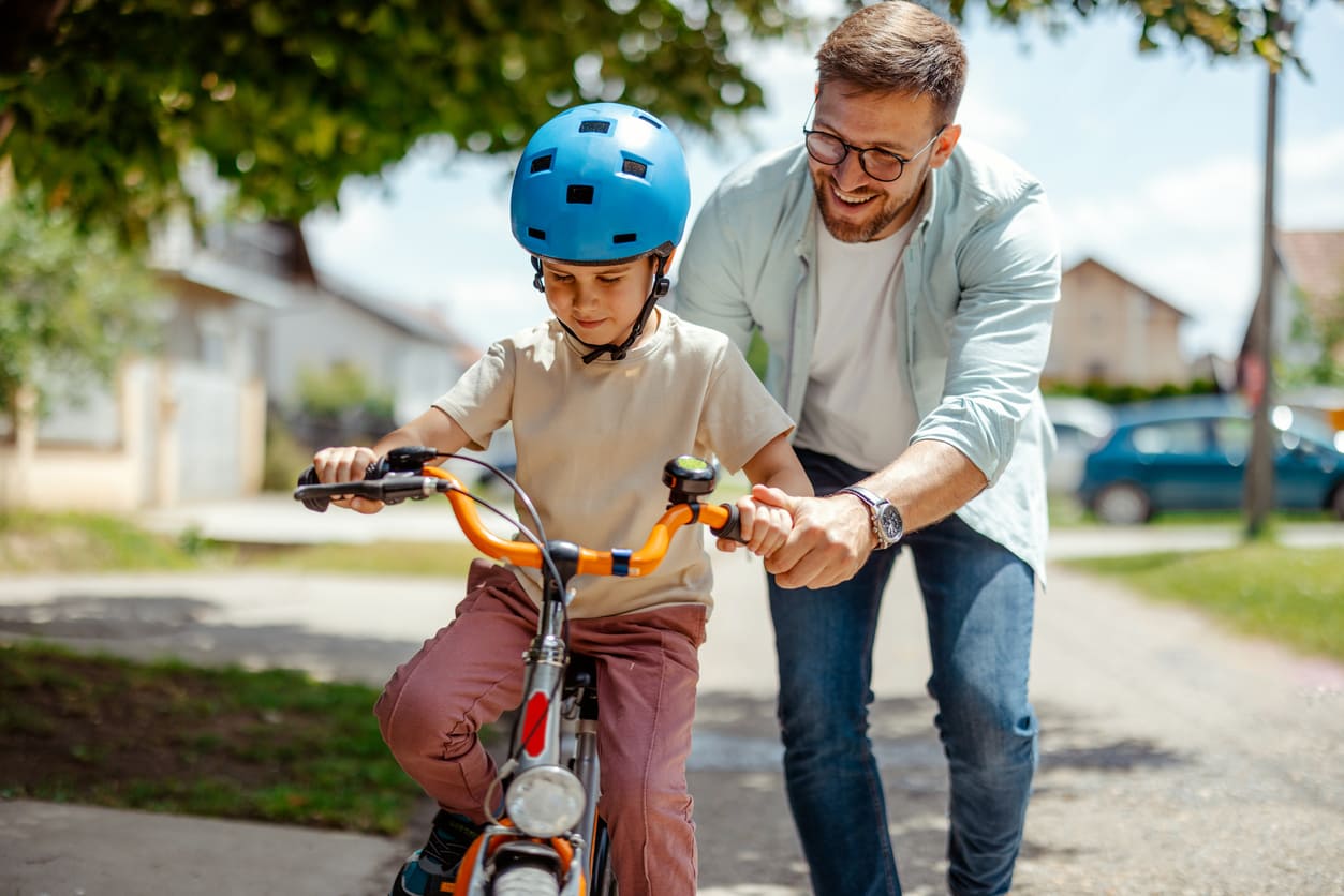 A smiling father helping his young child ride a bicycle, symbolizing support, care, and the everyday moments that define fatherhood.