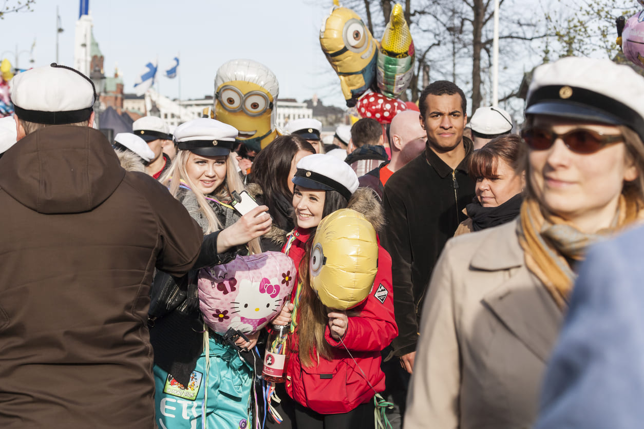 Cheerful students and families gathered outdoors, wearing white graduation caps and holding balloons, celebrating May Day in festive spring weather.