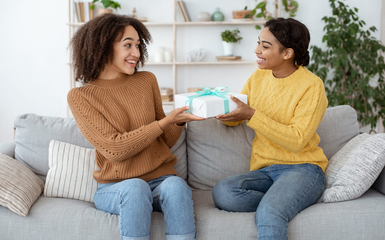 Two smiling friends sit on a sofa exchanging a wrapped gift, symbolizing warmth and appreciation on Ystävänpäivä in Finland.