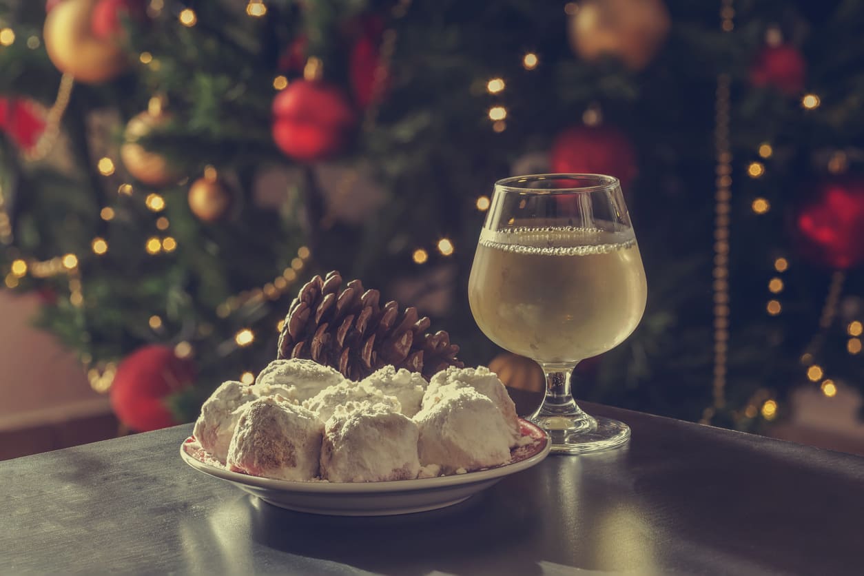 A plate of powdered kourabiedes and a glass of white wine sit beside a decorated Christmas tree, evoking the festive charm of a Greek Christmas.