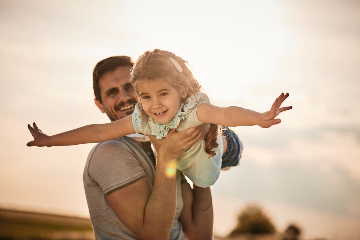 A smiling father holds his daughter high in the air at sunset, as she spreads her arms like wings in a playful, heartwarming moment.