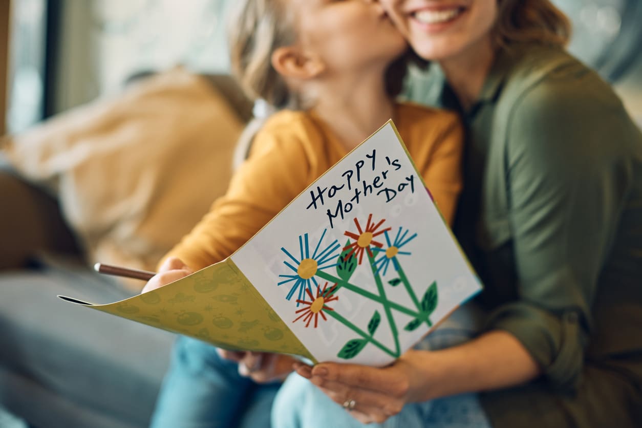 A smiling mother holds a colorful handmade card as her daughter kisses her cheek, capturing a joyful Mother's Day moment.
