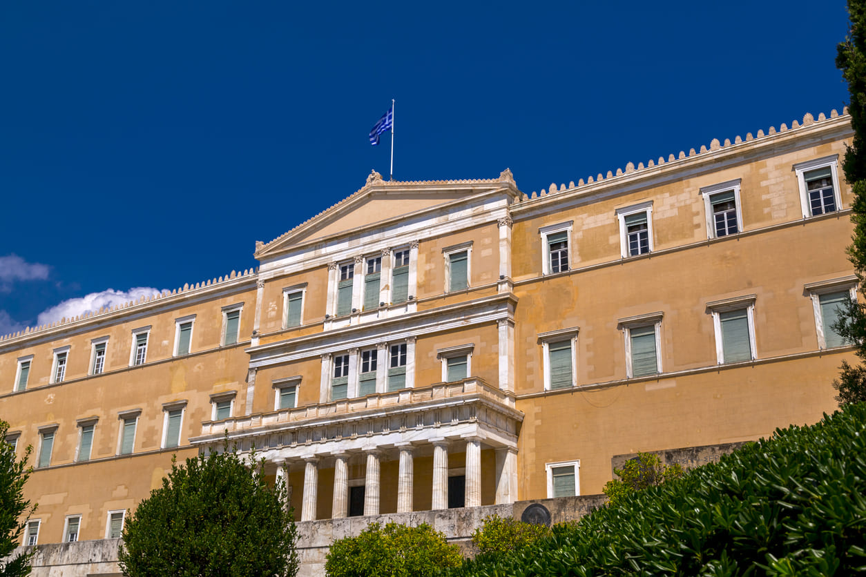 A bright view of the Hellenic Parliament building in Athens, crowned with the Greek flag under a clear blue sky, symbolizing national unity and democracy.