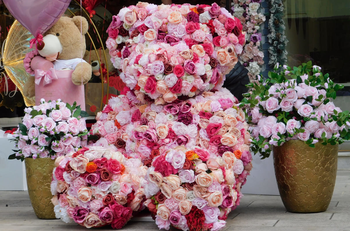 A storefront is decorated with a large teddy bear made of roses, surrounded by pink bouquets and heart-shaped balloons, creating a festive Valentine's Day display in Greece.