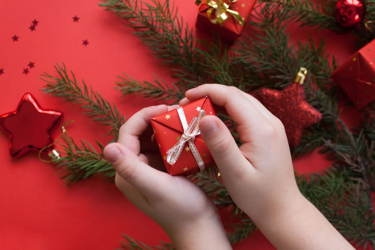 A close-up of a child's hands holding a small red gift box, surrounded by pine branches and festive decorations, evoking the joy of Christmas Eve.