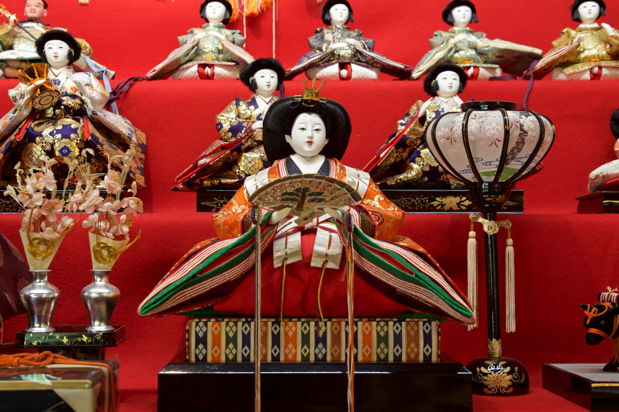 A tiered display of beautifully dressed hina dolls stands in a warmly lit room, surrounded by festive decorations and family offerings.