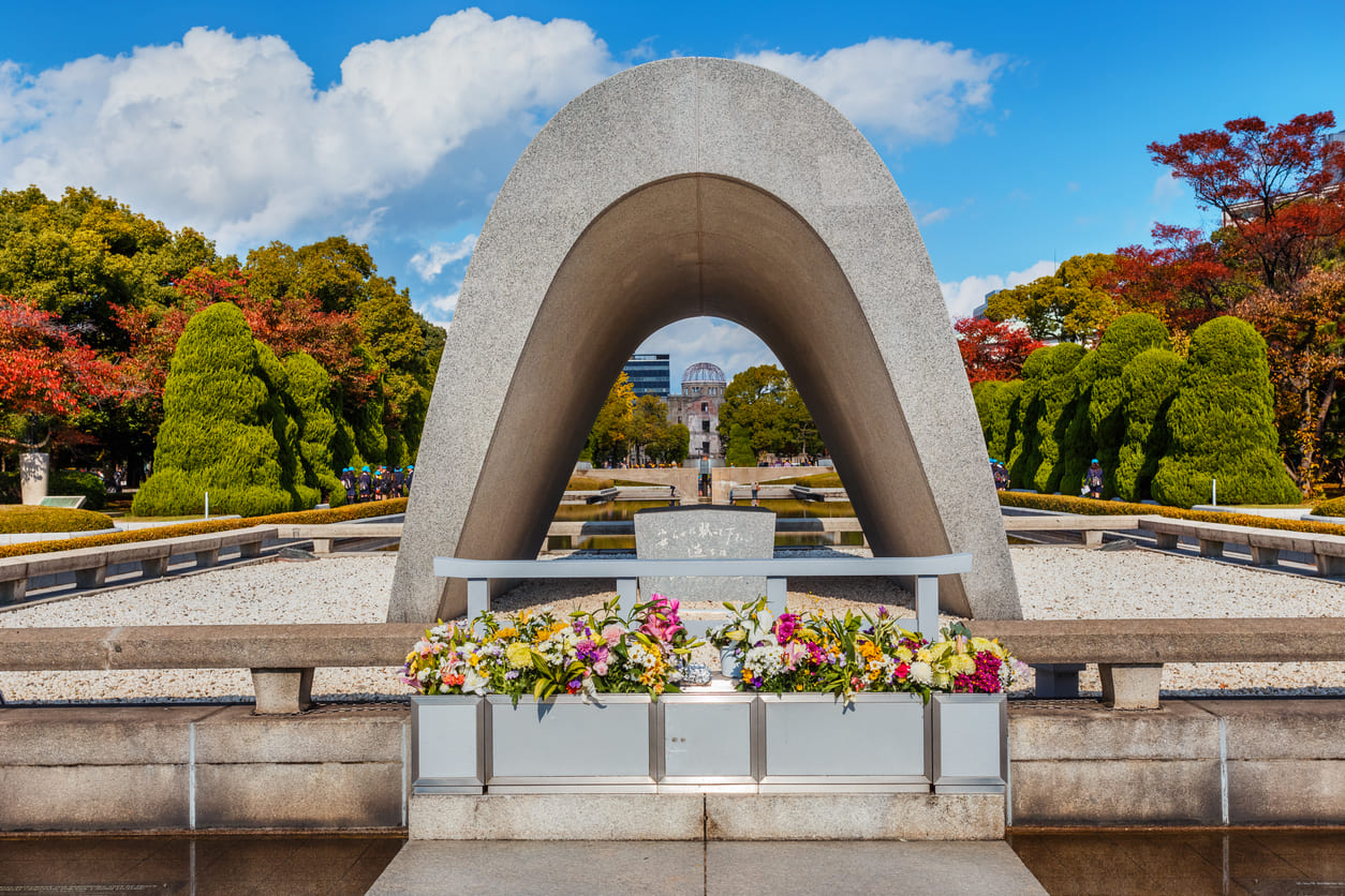 A solemn stone arch at Hiroshima Peace Memorial Park is adorned with flowers, framing the Atomic Bomb Dome in the distance beneath a clear blue sky.