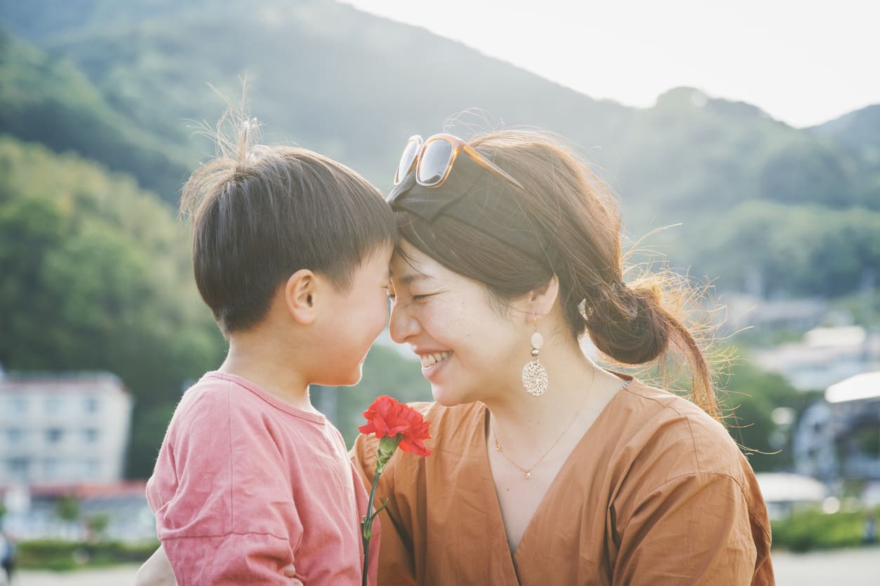 A young boy and his mother share a touching moment outdoors, smiling warmly as he holds a red carnation in his hand.