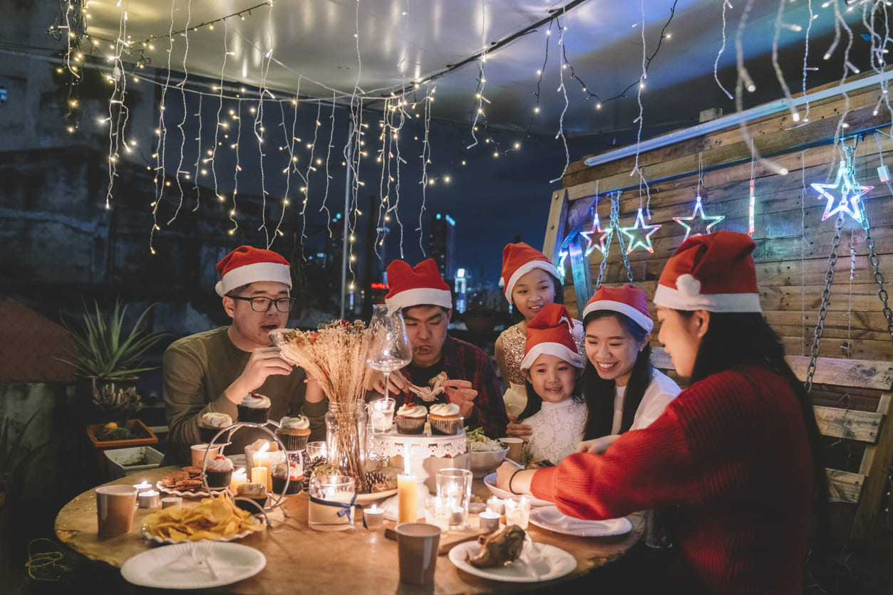 A Malaysian family wearing Santa hats, gathered around a candlelit table under festive lights, enjoying a joyful Christmas celebration together.