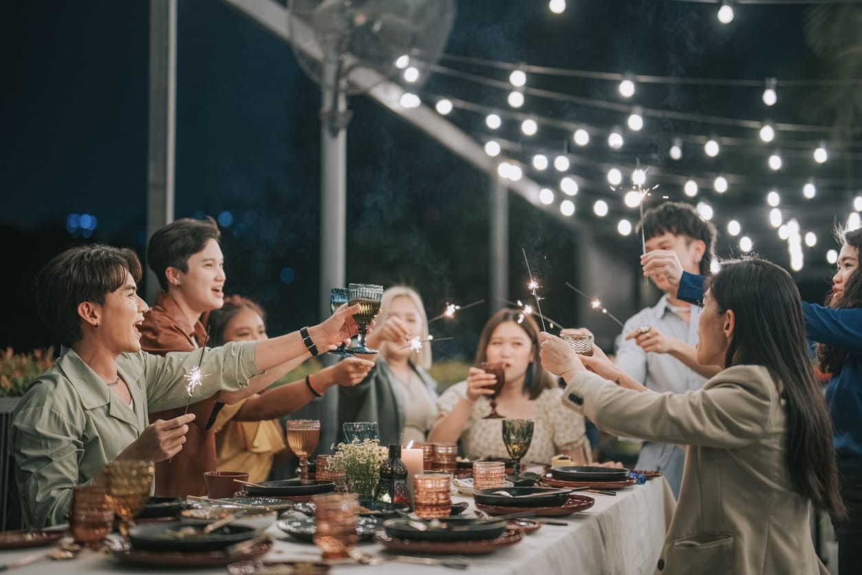 A group of friends gathered around a decorated dinner table, holding sparklers and toasting under string lights in a joyful New Year's Eve celebration.
