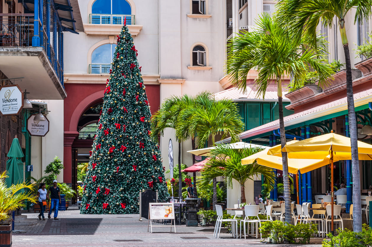 A tall, festively decorated Christmas tree surrounded by palm trees, shops, and people enjoying the holiday atmosphere in Port Louis, Mauritius.