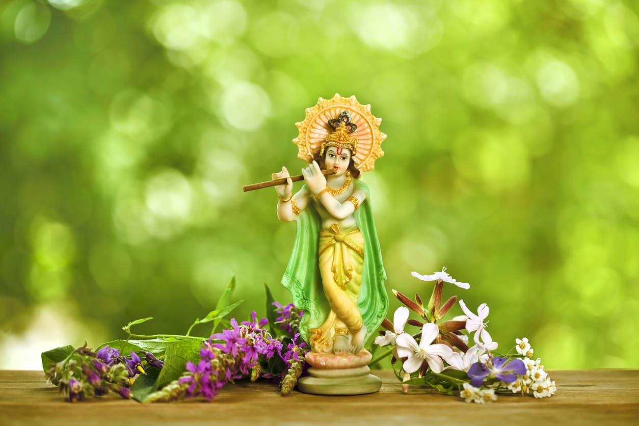 A vibrant idol of Lord Krishna with a flute, surrounded by fresh flowers, set against a soft green background, symbolizing the festive spirit of Janmashtami.