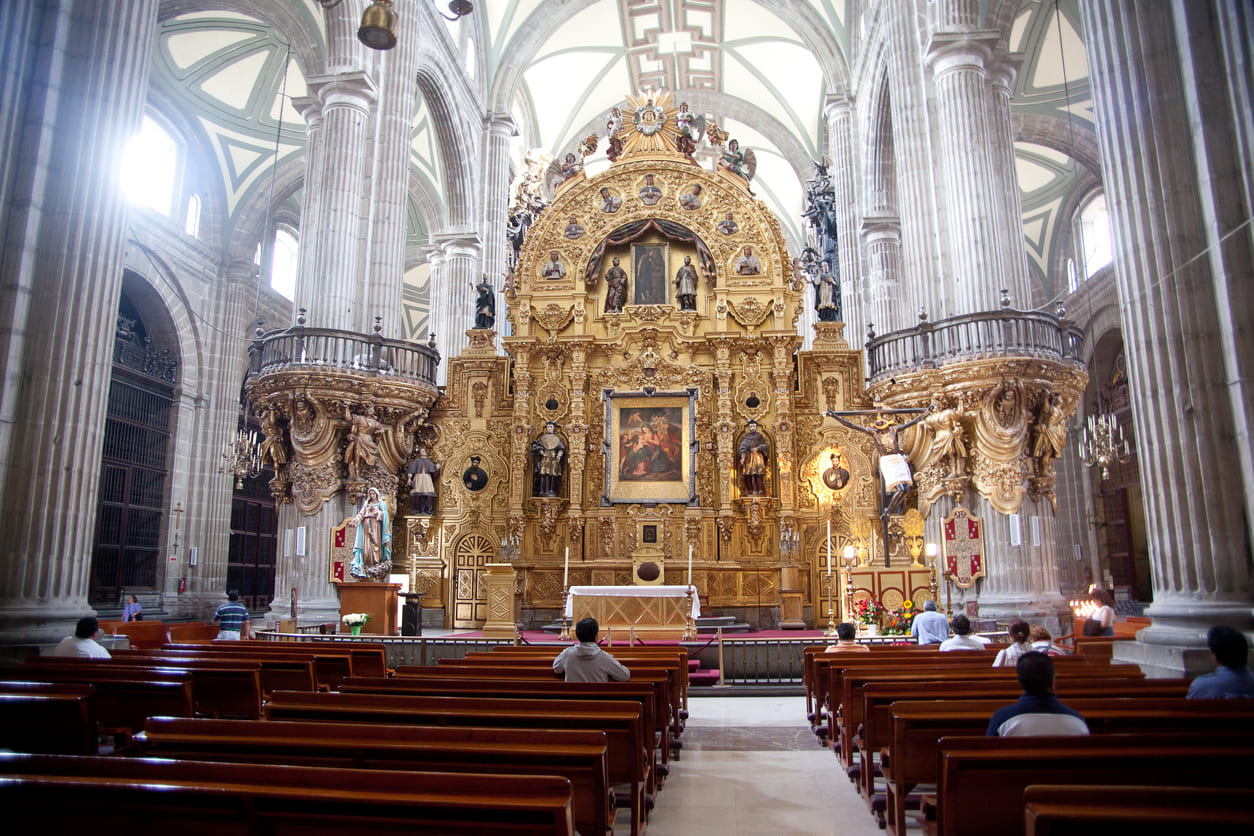 The grand interior of the Metropolitan Cathedral in Mexico City, where faithful sit in prayer beneath ornate arches and religious artwork.