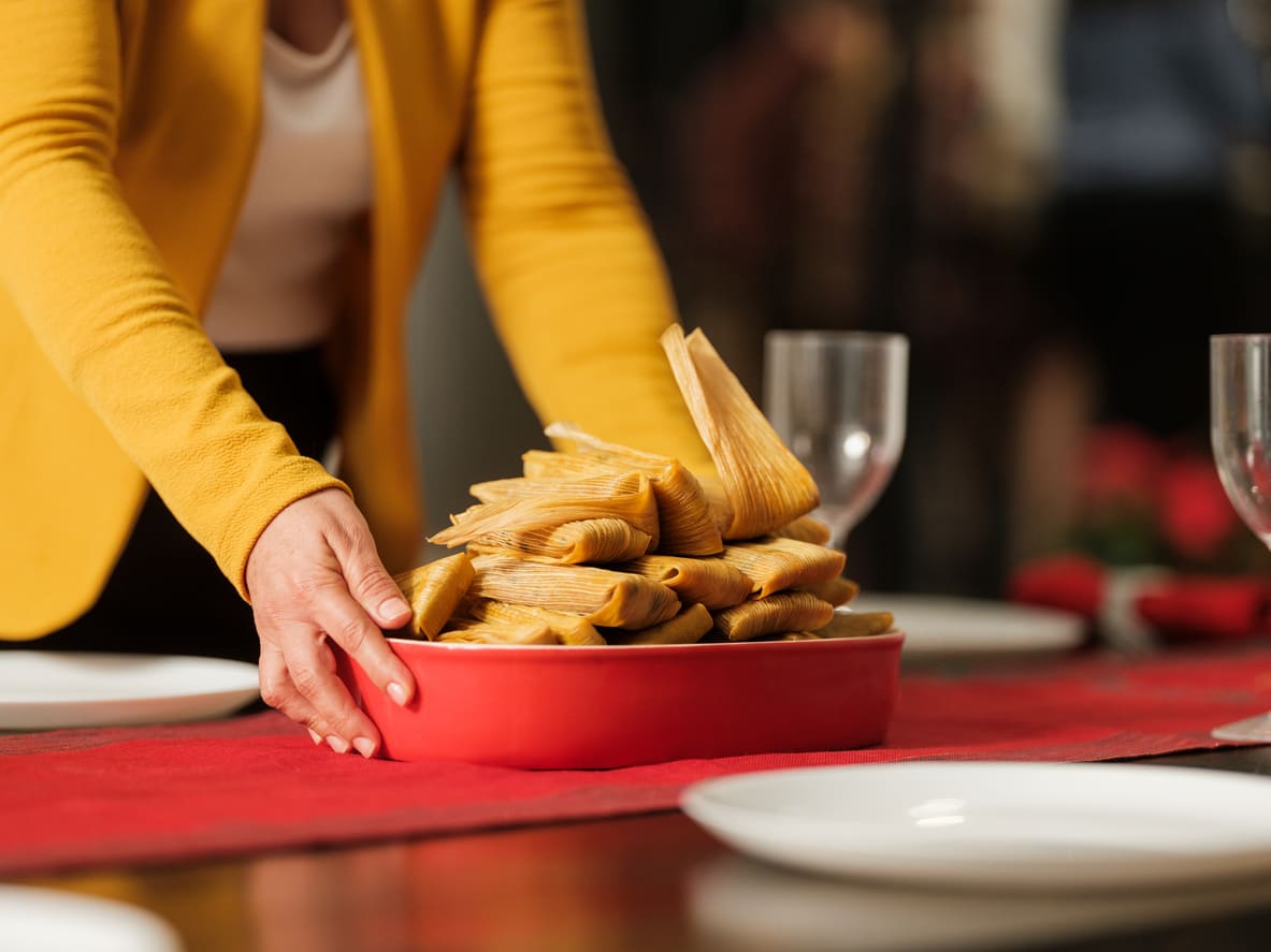 A person placing a festive bowl of tamales on a set dining table, capturing a key tradition of Candlemas in Mexico.