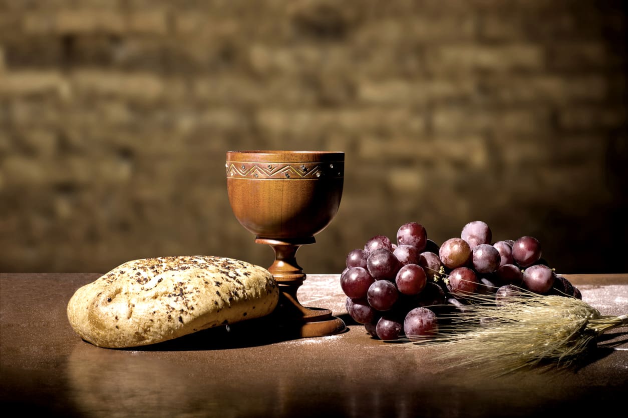 A rustic setup with a loaf of bread, a wooden chalice, fresh grapes, and wheat, symbolizing the elements of the Holy Eucharist.