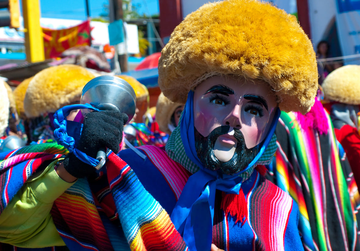 A person wearing a colorful traditional outfit and a painted mask, participating in a lively street celebration in Mexico.
