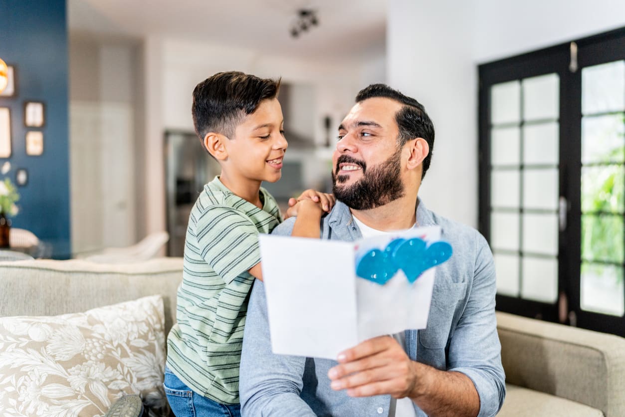 A father beams with pride as his son hands him a colorful handmade Father's Day card.