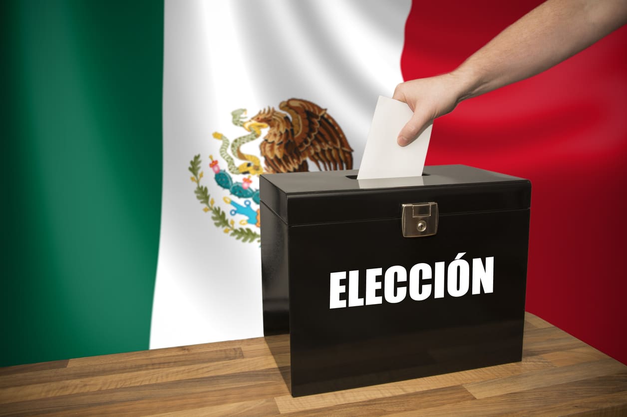 A hand placing a ballot into a black box, with the Mexican flag in the background, symbolizing national voting.