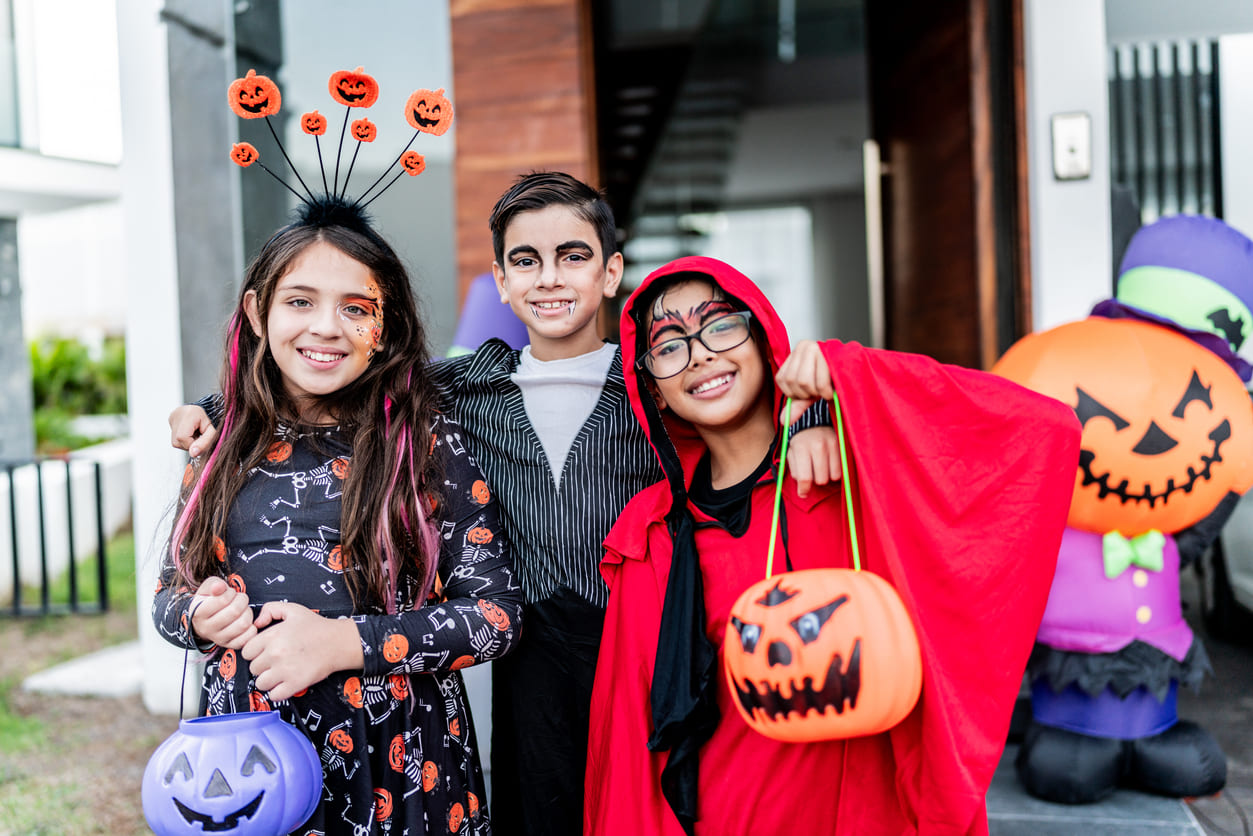 Three children in colorful Halloween costumes, holding pumpkin buckets and smiling outside a decorated home.