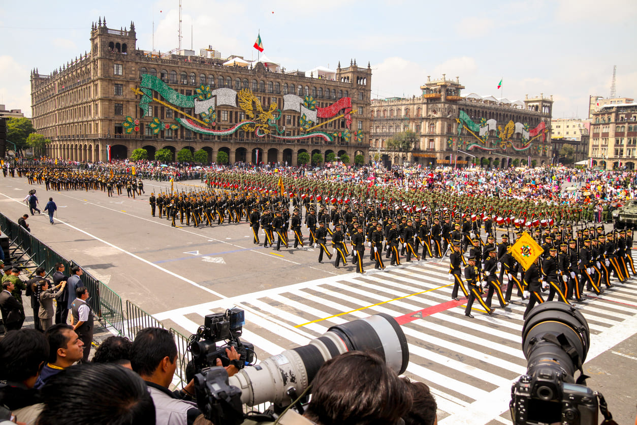 The image shows a grand military parade in Mexico City's Zócalo, with crowds watching and buildings decorated in national colors.
