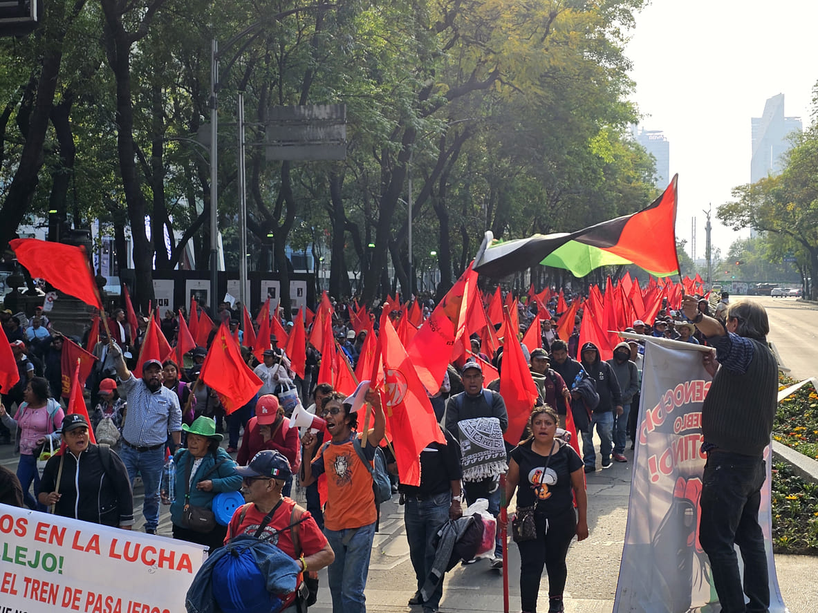 A large group of people marching through a tree-lined avenue with red flags and banners, demonstrating in support of workers' rights on Labor Day in Mexico.