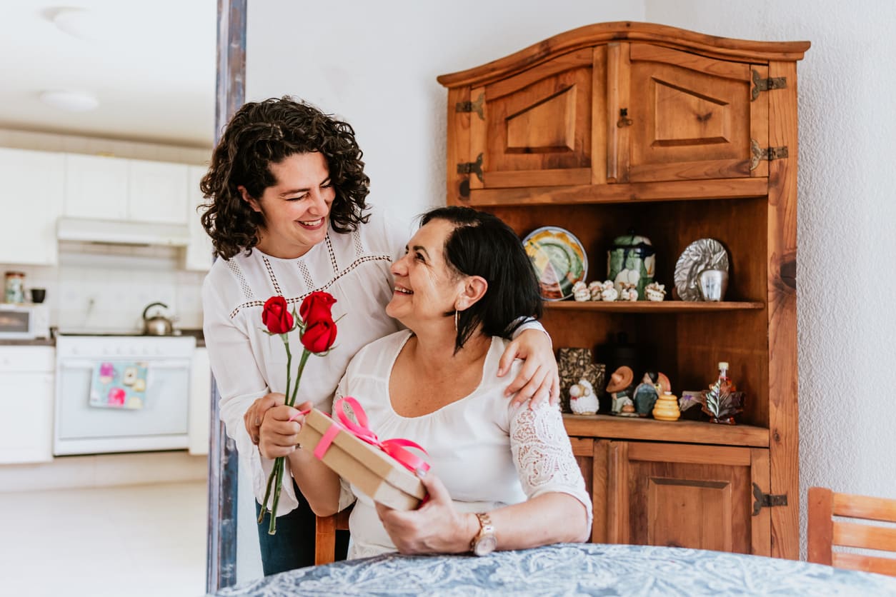 A smiling daughter gives her mother red roses and a gift while they share a warm moment at home.