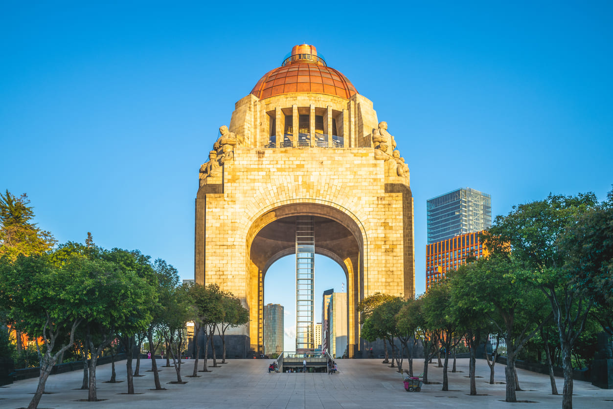 The Monument to the Revolution in Mexico City, a grand stone structure with a domed roof, surrounded by trees and modern buildings.