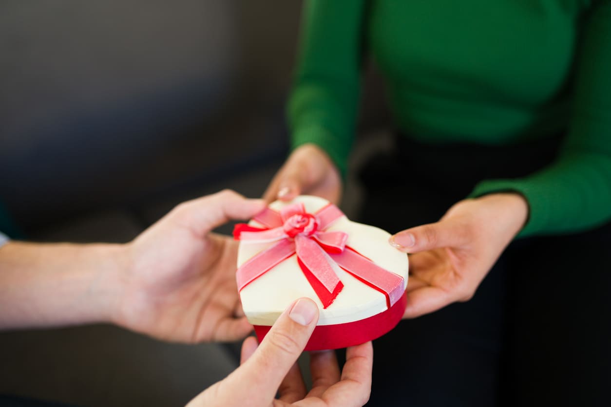 Two people exchanging a heart-shaped gift box with a pink ribbon, symbolizing affection and celebration on Valentine's Day.