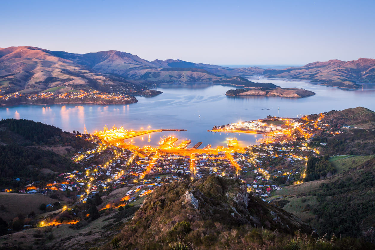 An illuminated view of Lyttelton Harbour at dusk, surrounded by rolling hills, marking the historic gateway of Canterbury's early European settlers.