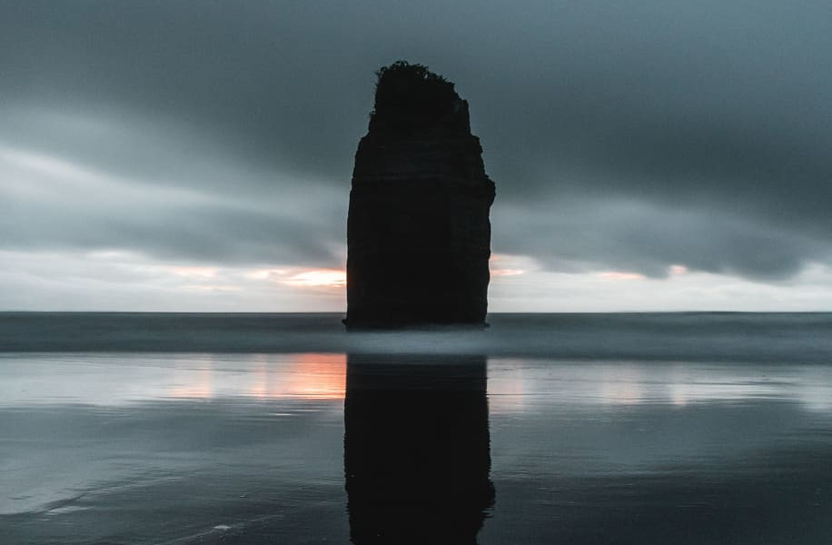 A solitary sea stack from the Three Sisters rock formation rises against a moody sky, reflected in the calm waters of Cape Pattisson on Chatham Island.