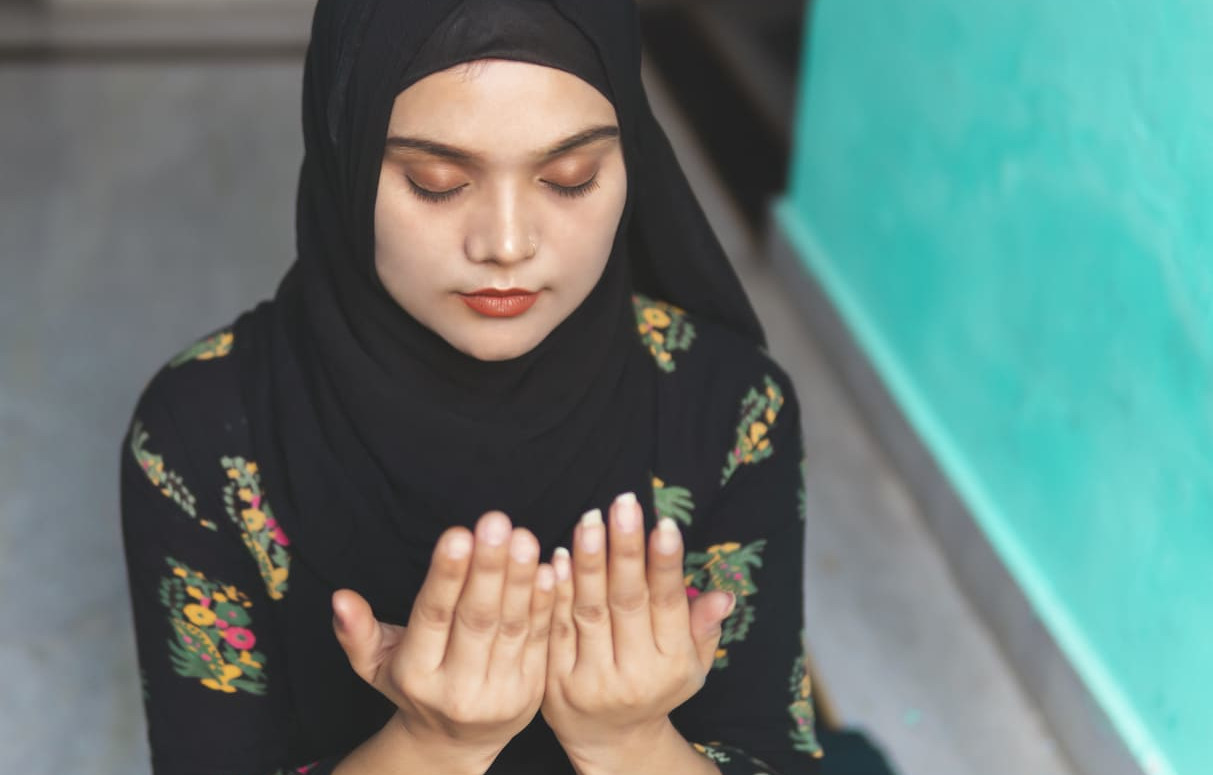 A Muslim woman in a black hijab praying with cupped hands, reflecting the solemn observance of Maulid un-Nabi.