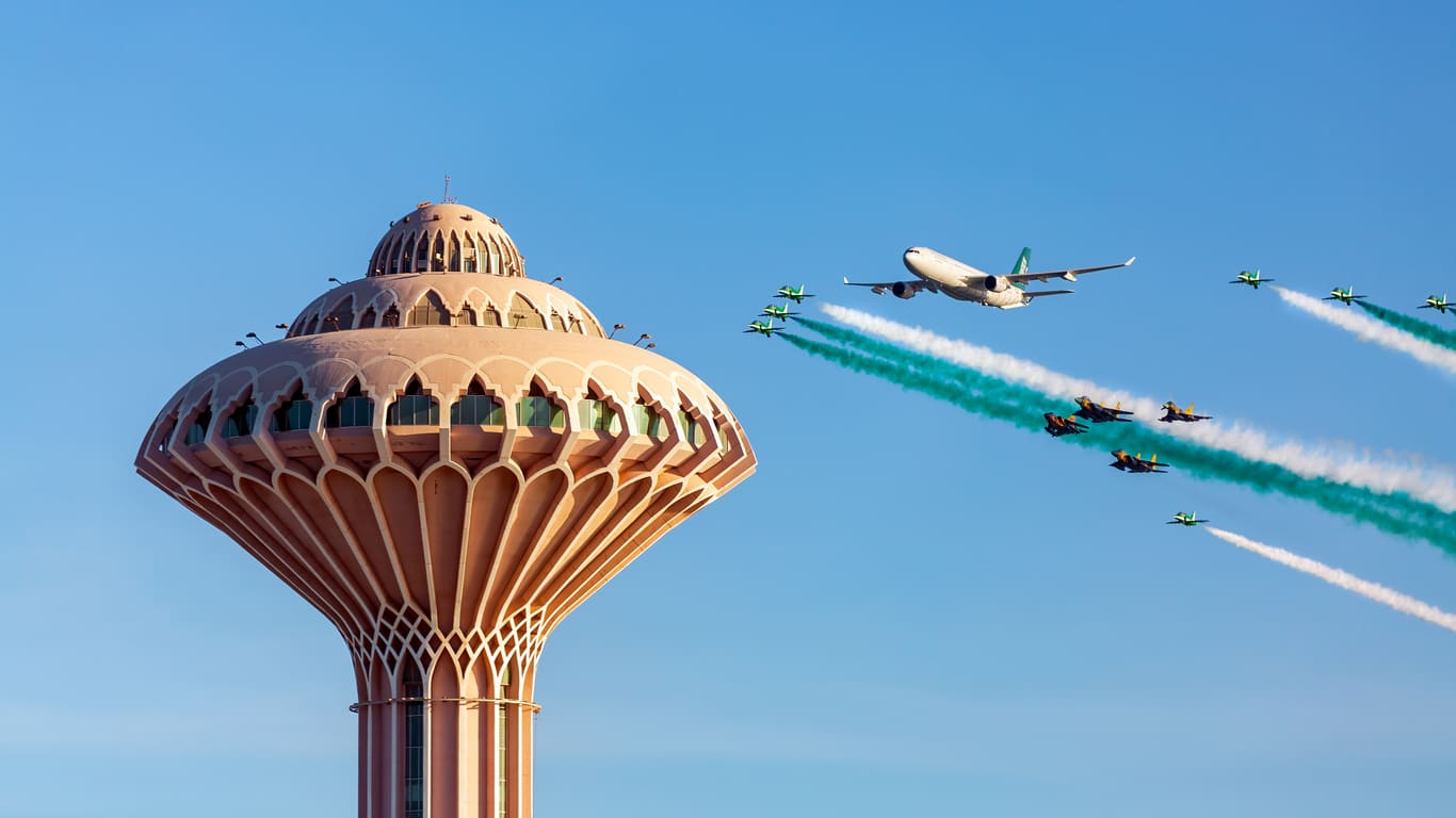 A formation of Saudi jets and an aircraft perform a vibrant air show near Al Khobar's iconic water tower, celebrating national pride.