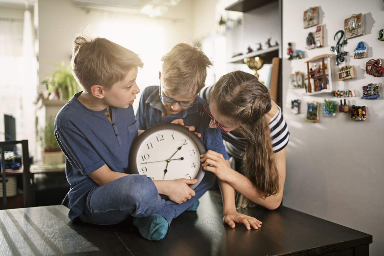 Smiling children adjusting a wall clock, playfully plotting a prank to surprise their parents on April Fool's Day.