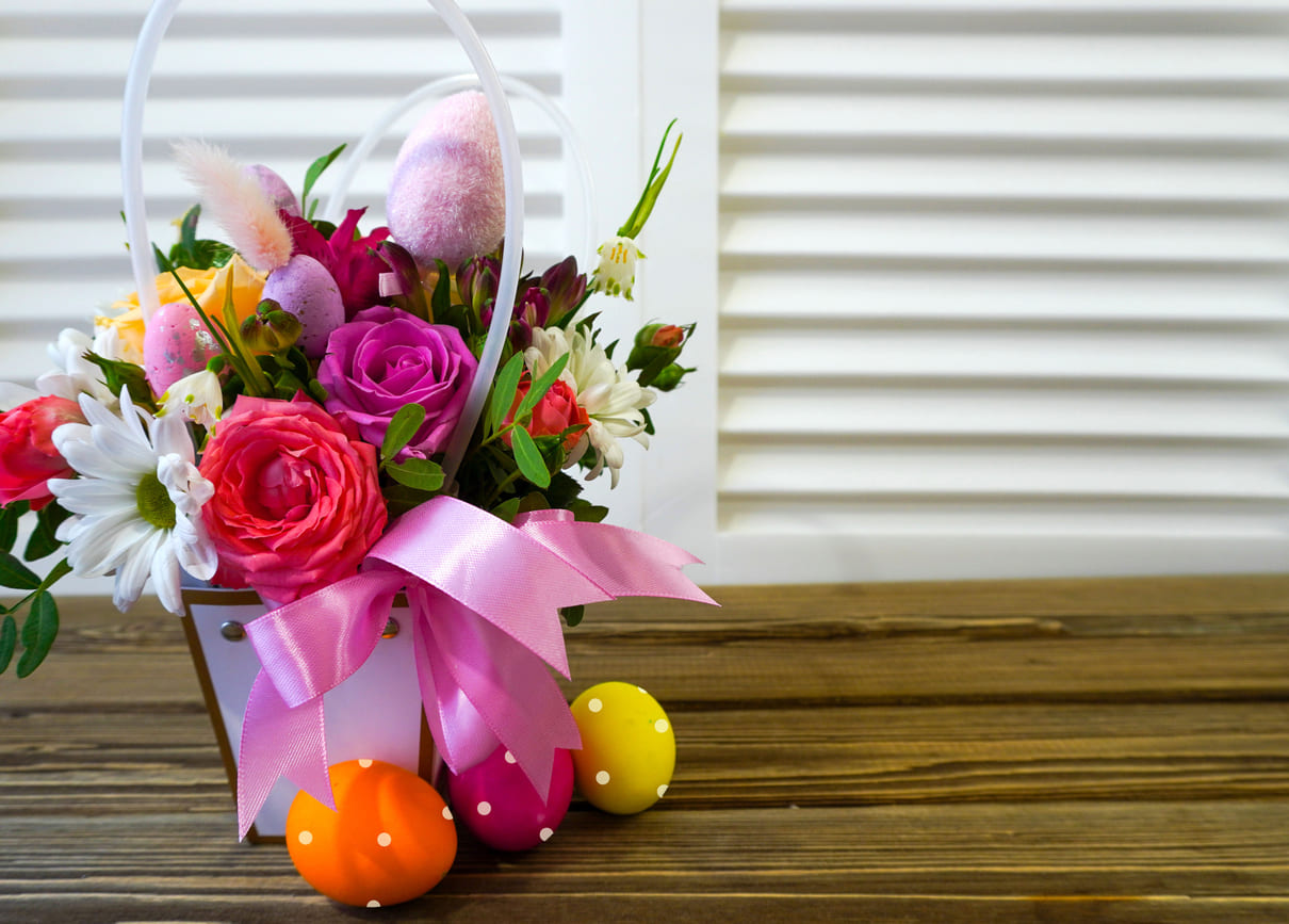 A vibrant Easter basket filled with colorful flowers and polka-dotted eggs, set on a wooden table against a white backdrop.
