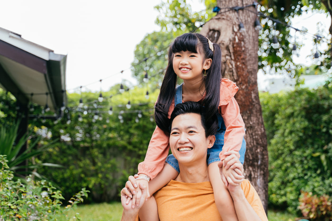 A smiling father carrying his happy daughter on his shoulders in a lush garden setting, reflecting warmth, bonding, and playful joy.