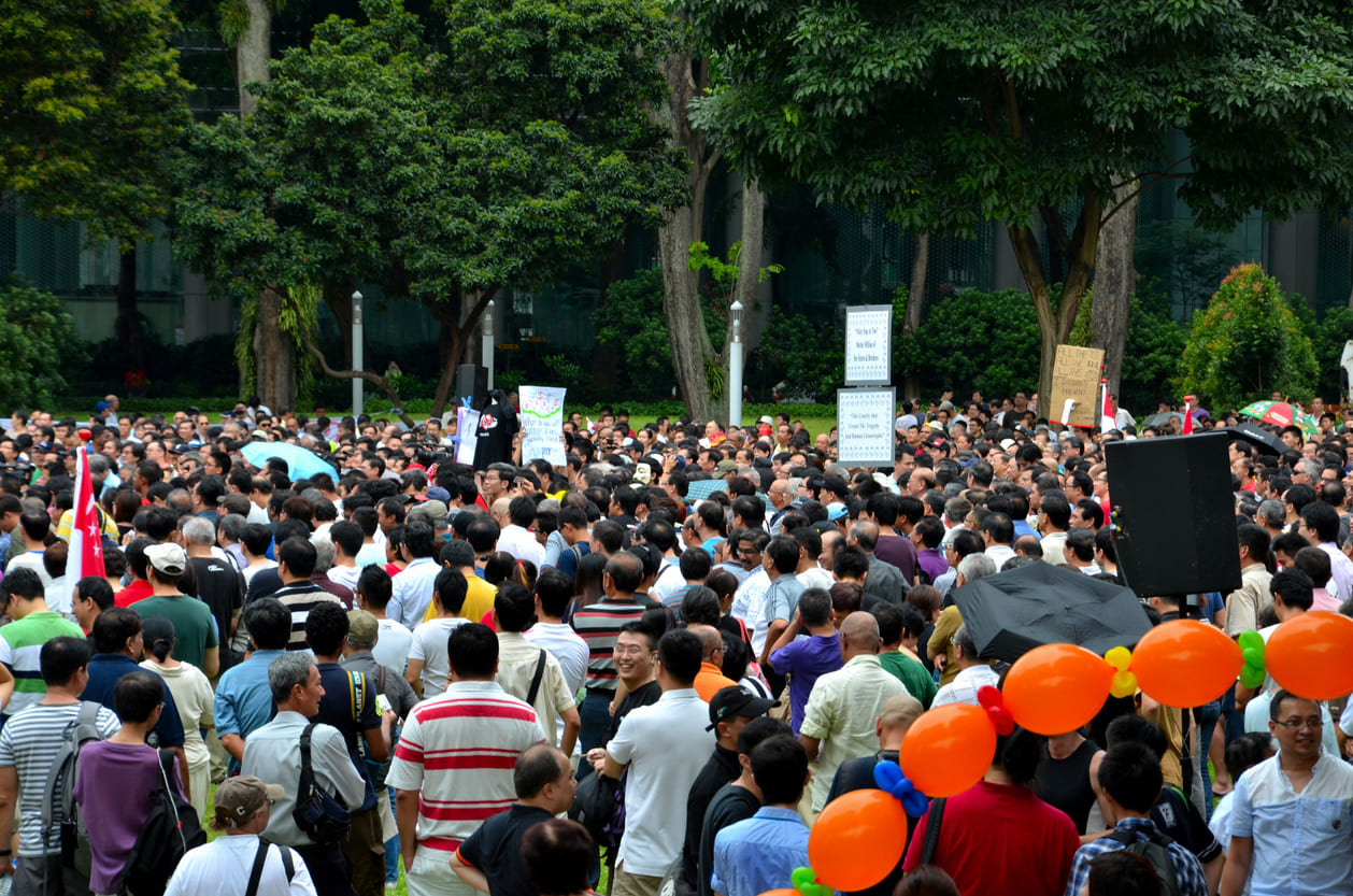 A large crowd gathered outdoors with signs and balloons, capturing the energy and solidarity of a May Day rally in Singapore.