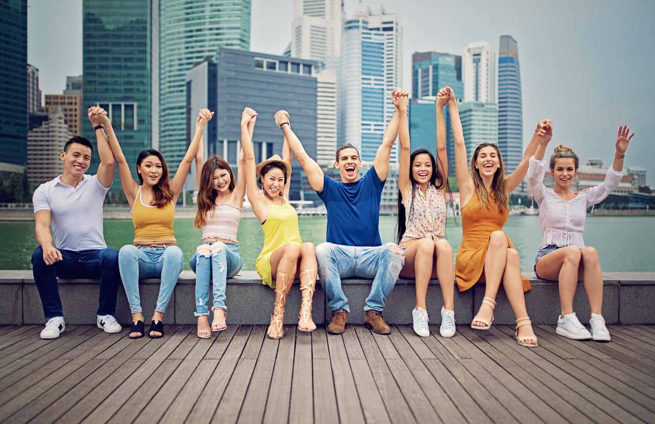 A diverse group of smiling youths holding hands with the Singapore skyline in the background, representing harmony and togetherness.