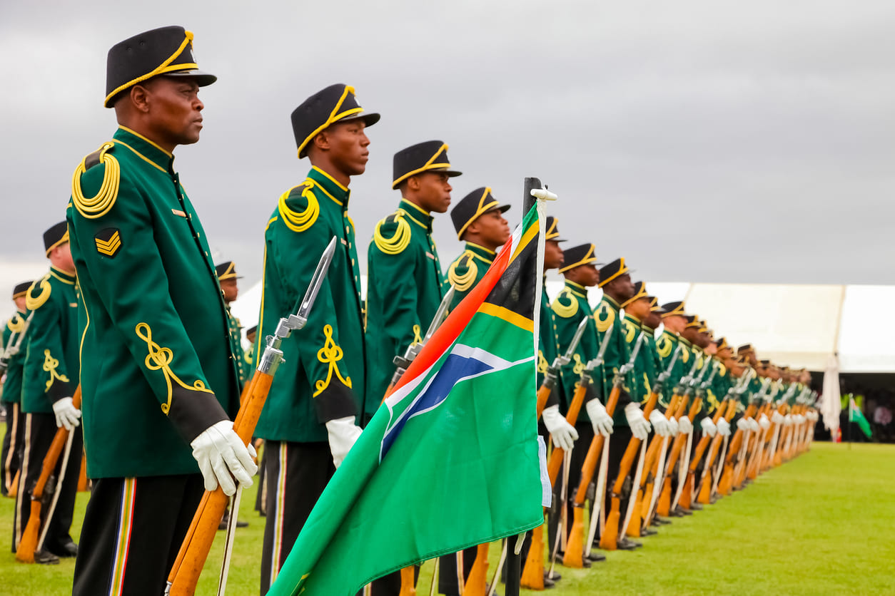 A line of South African soldiers in formal green uniforms standing in formation with rifles and the national flag during a ceremonial Armed Forces Day event.