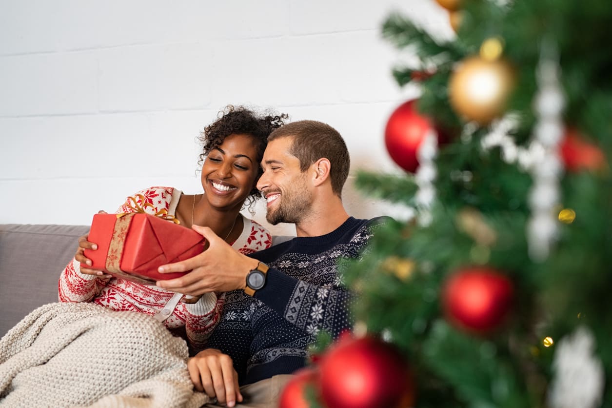 A smiling couple sitting together on a couch, exchanging a wrapped gift beside a beautifully decorated Christmas tree.