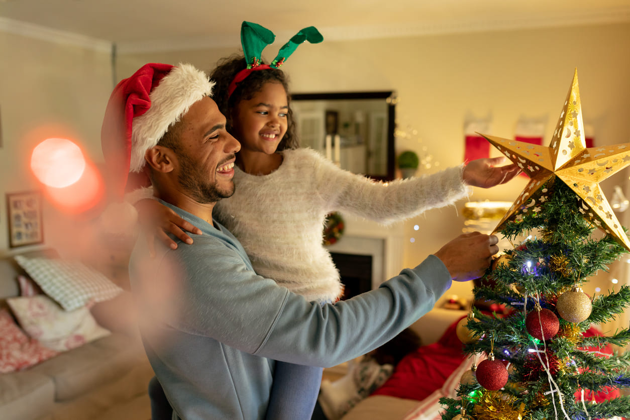 A smiling father holding his daughter as she places a golden star on a brightly decorated Christmas tree, creating a heartwarming festive scene.