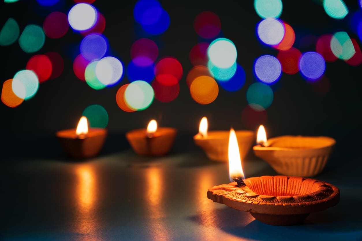 A row of traditional Diwali oil lamps shines brightly against a backdrop of colorful festive lights.