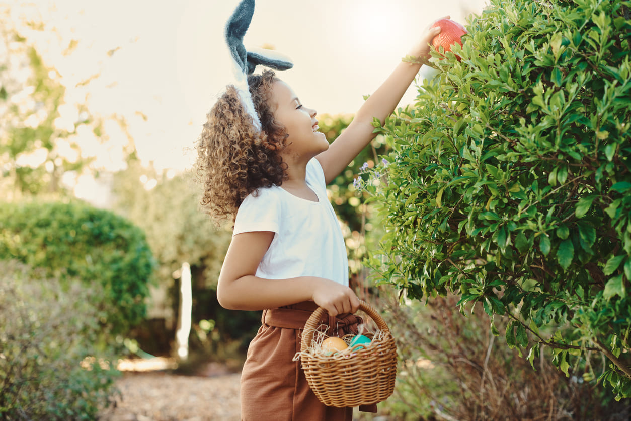 A smiling child wearing bunny ears, reaching into a bush with a basket of Easter eggs, enjoying a festive outdoor egg hunt.