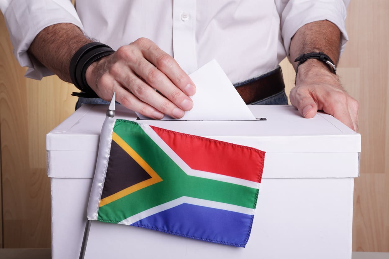 A close-up of a person placing a ballot into a voting box decorated with the South African flag, capturing the act of democratic participation.
