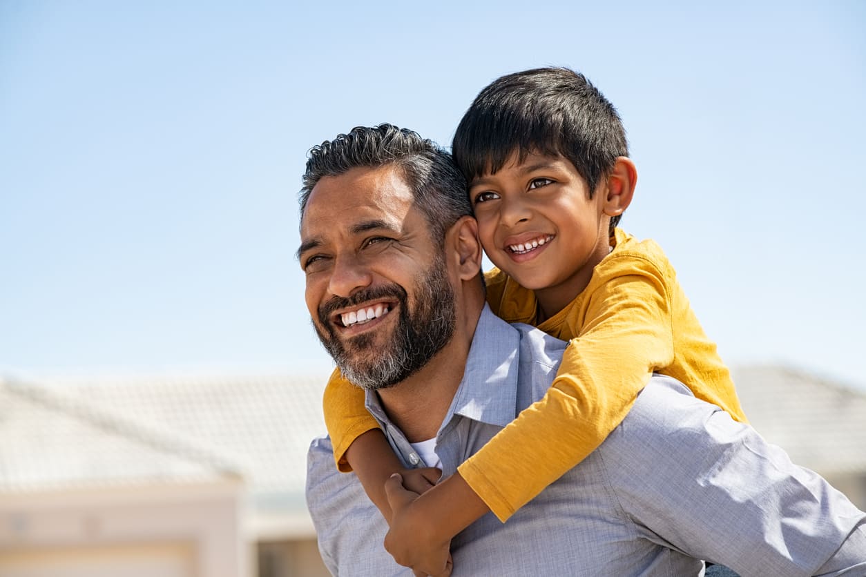 A smiling father carries his happy son on his back, enjoying a bright and heartwarming moment outdoors.