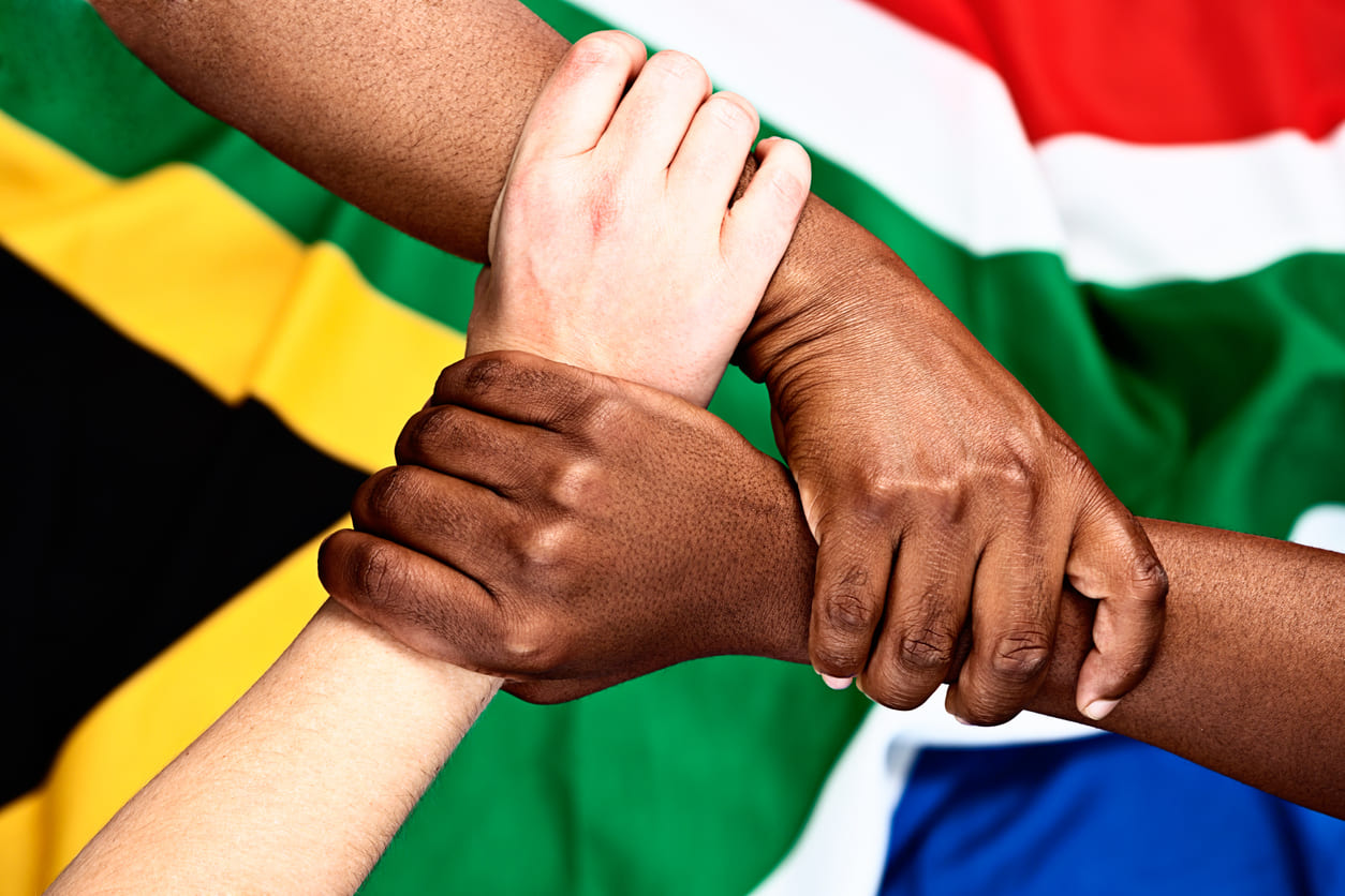 Hands of different skin tones firmly holding each other in front of the South African flag, symbolizing unity, solidarity, and shared human rights.