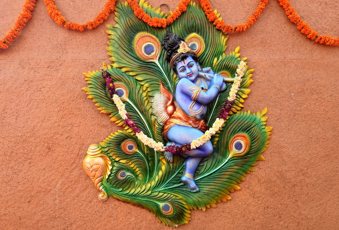 A colorful wall decoration of baby Krishna seated on peacock feathers, adorned with garlands and festive flowers.